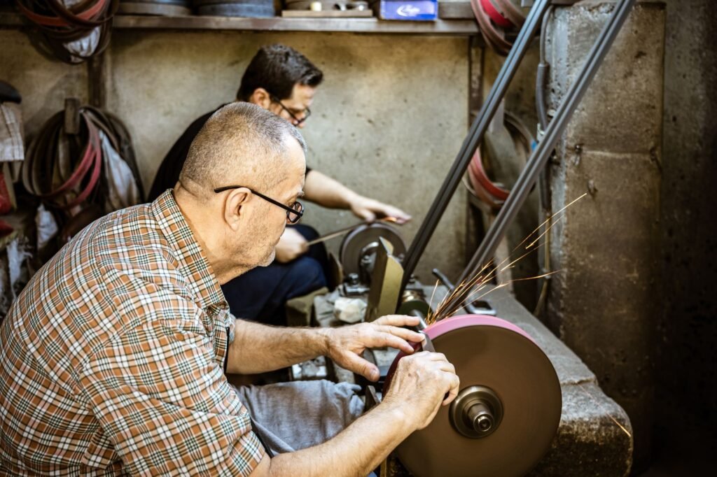 Two skilled blacksmiths sharpening metal tools on a grinding wheel indoors.